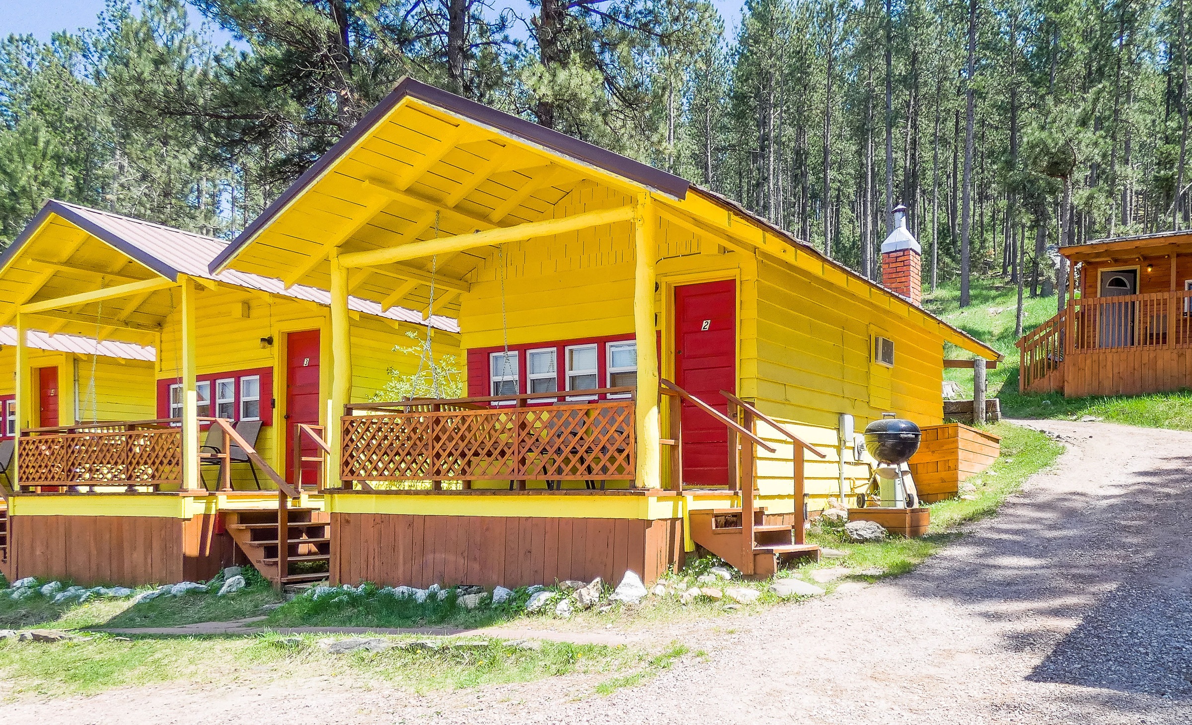 Cabin 2 seen from the side: yellow with a red door, porch rail and a charcoal grill
