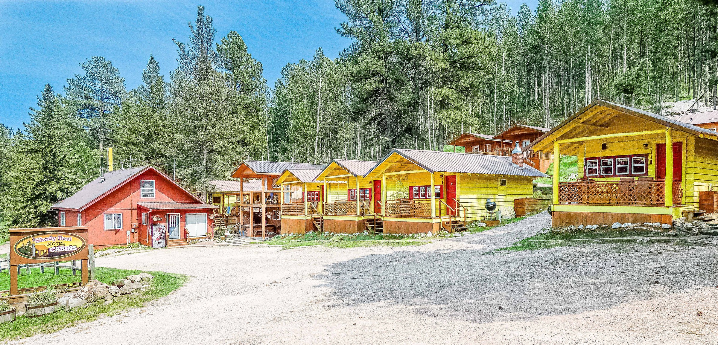 Yellow cabins with red doors and porches in a row beside the red office and the property sign, beneath a pine-covered hillside